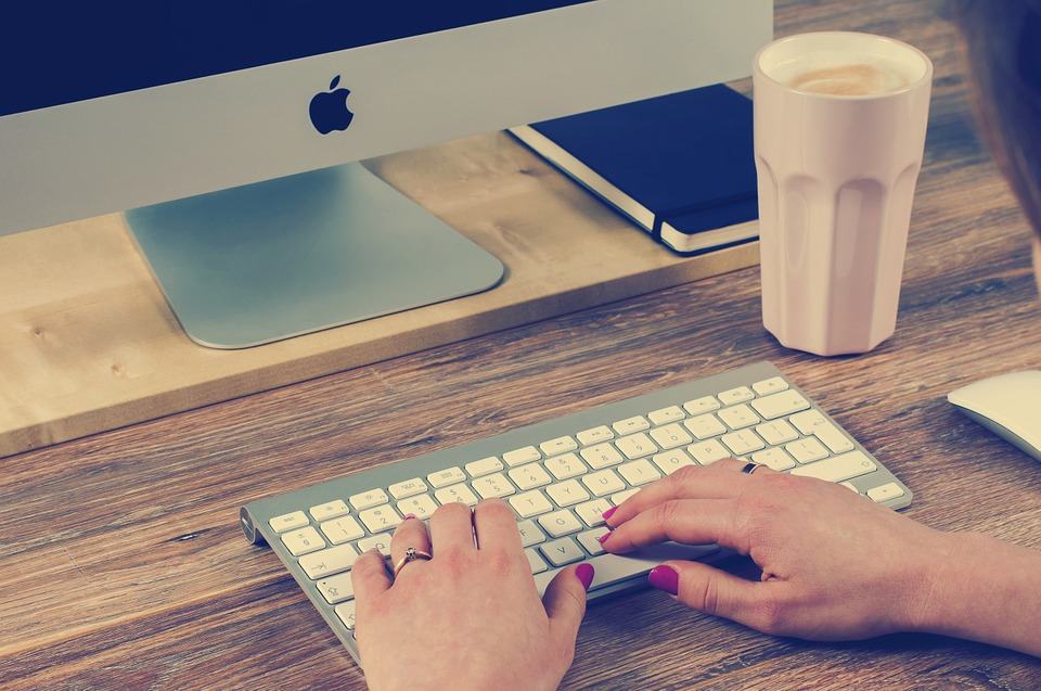Woman typing on an Apple iMac keyboard with a coffee cup beside her, representing local business content creation