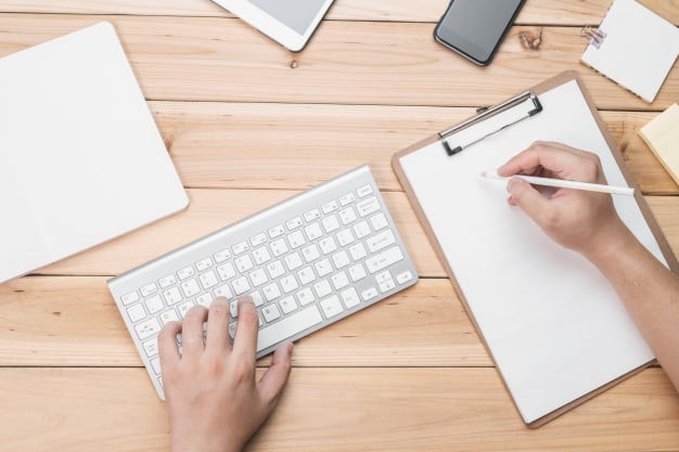 Overhead view of hands typing on a keyboard while writing notes on a clipboard, representing guest post content creation
