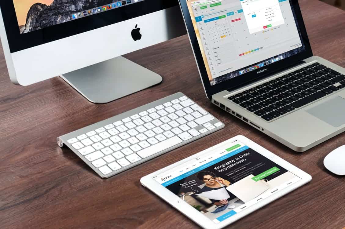 iMac, MacBook Pro and iPad on a wooden desk displaying website and app interfaces