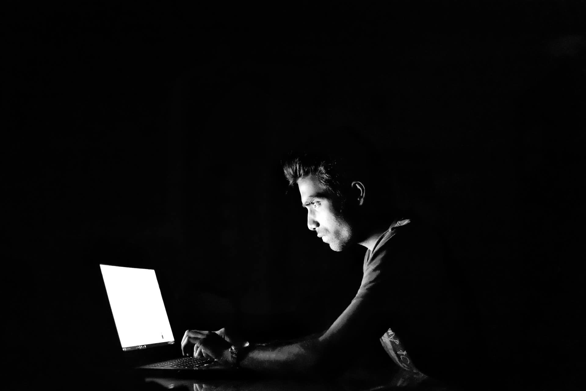 Man typing on a laptop in a dark room, lit only by the screen glow, illustrating hacking or cybersecurity
