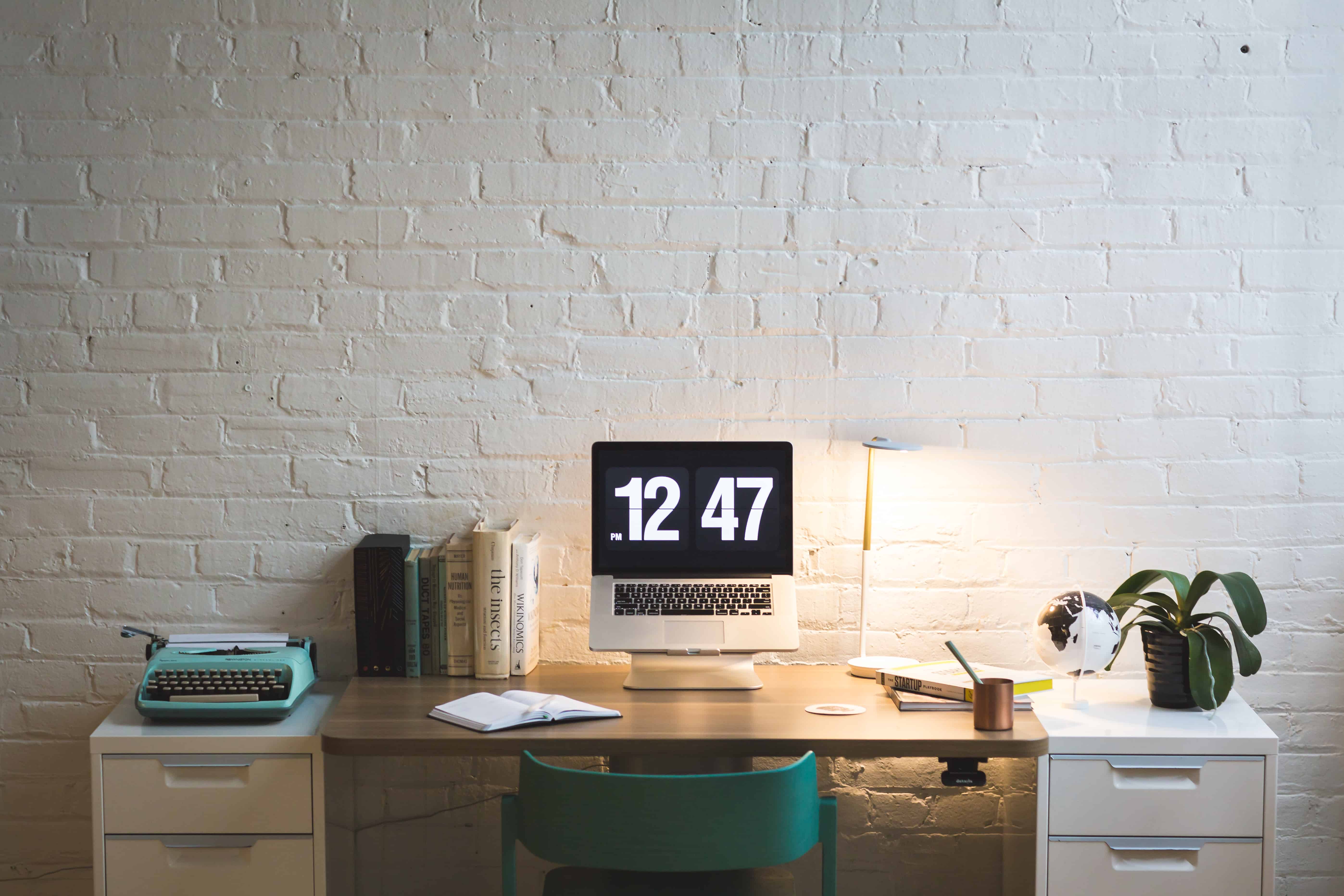 Minimalist home office desk with iMac clock display, typewriter, books, and lamp against a white brick wall