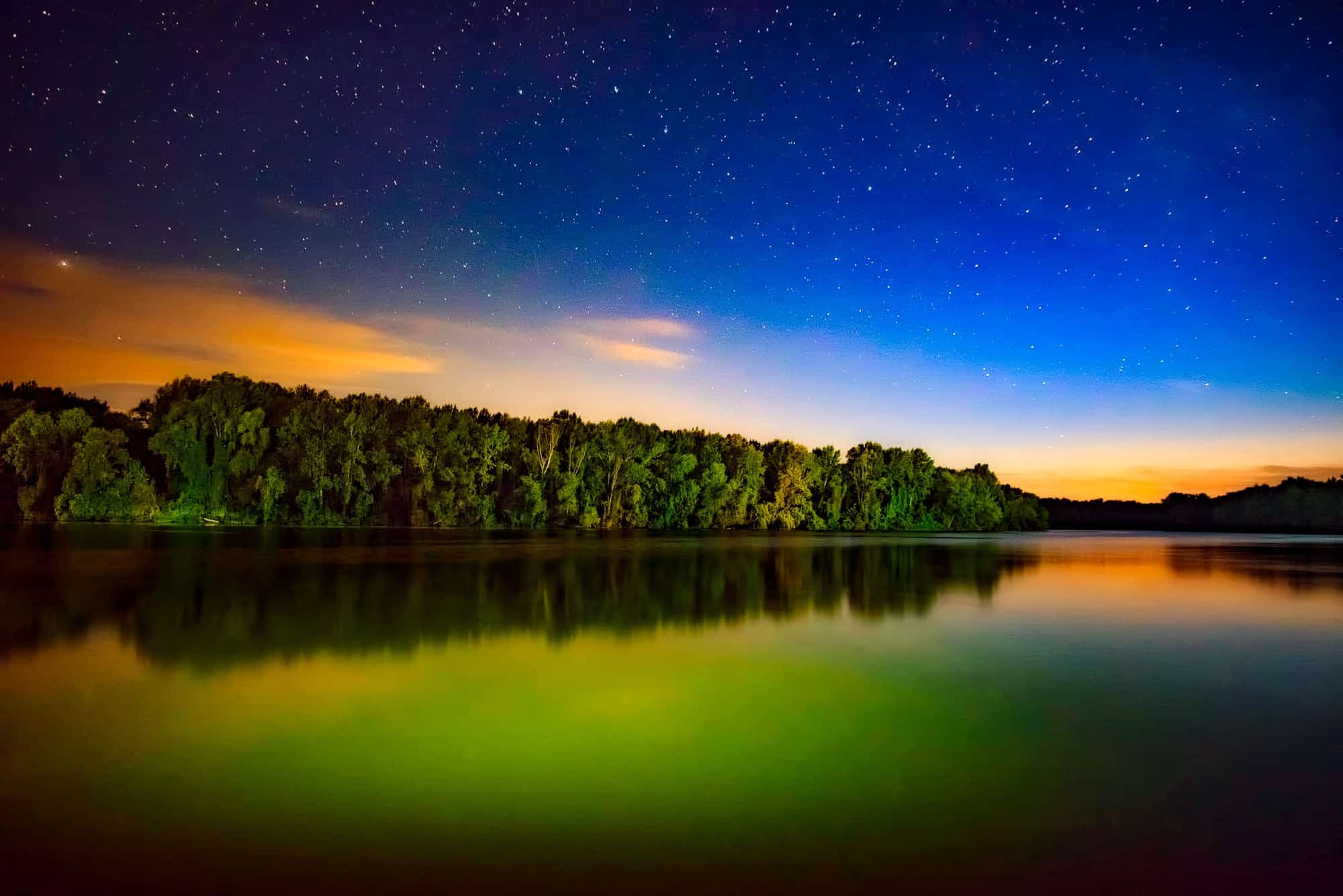 Vivid night-sky landscape over a lake with starry blue sky and orange twilight glow reflecting in the water