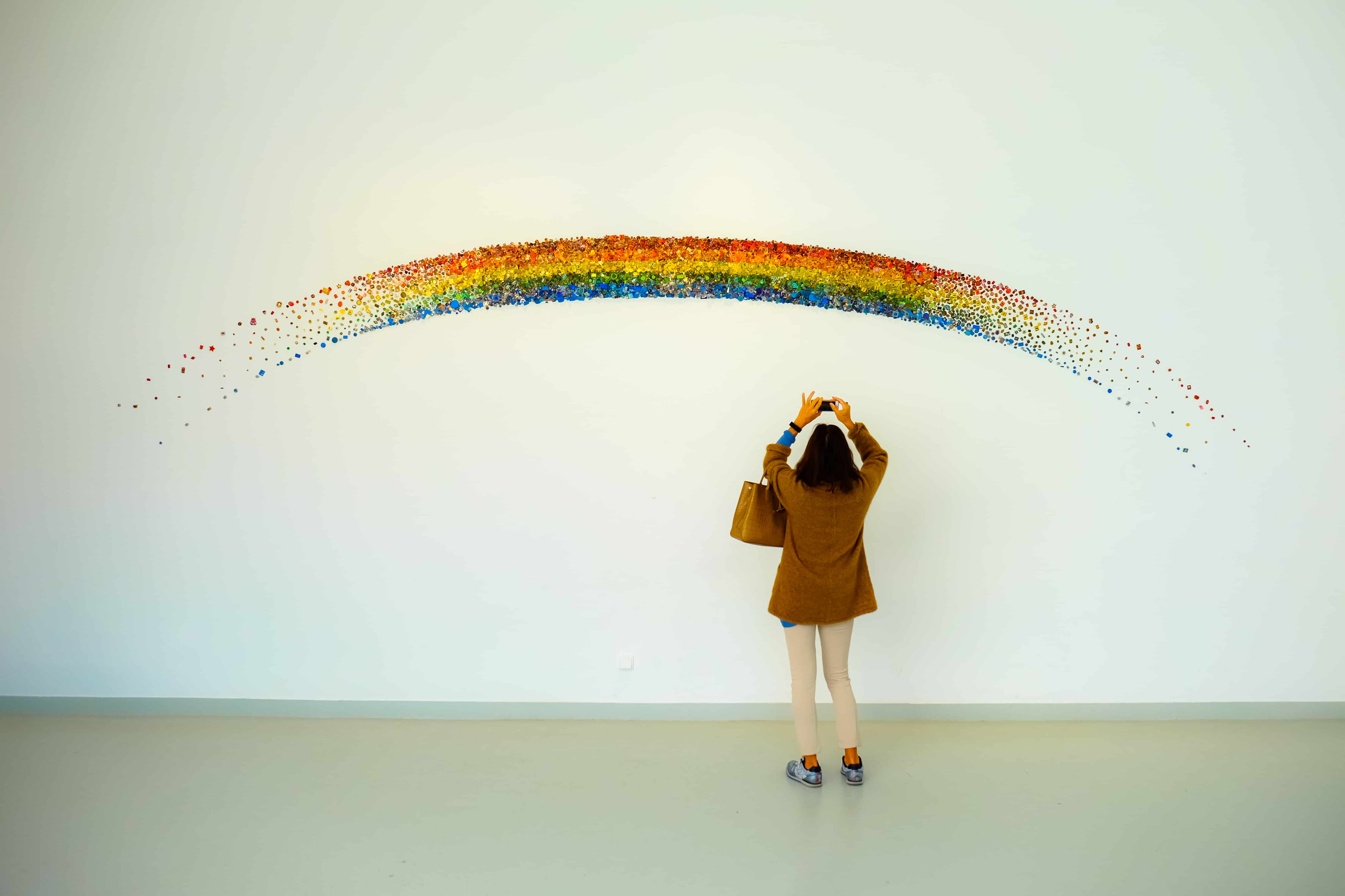 Gallery visitor photographing a rainbow artwork made of coloured dots mounted on a white wall