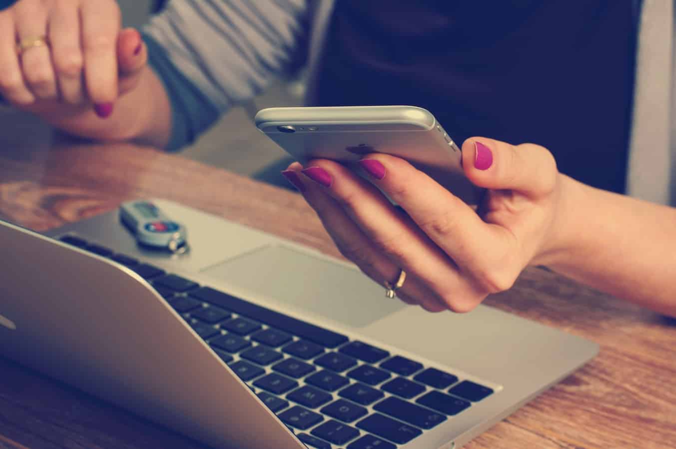 Close-up of a woman holding an iPhone above a laptop keyboard on a wooden desk