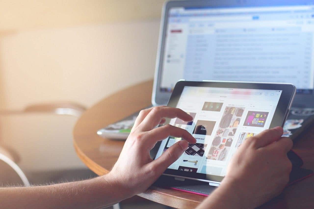 Hands browsing a shopping website on a tablet with a laptop and email inbox visible in the background