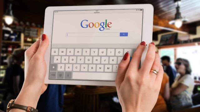 Woman typing on a tablet displaying the Google search homepage with an on-screen keyboard in a cafe setting