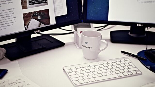 Designer's desk with dual Dell monitors showing website layouts, Apple keyboard, Twitter mug, and notebook