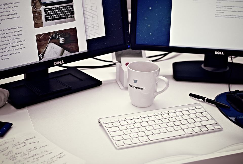 Designer's desk with dual Dell monitors showing website layouts, Apple keyboard, Twitter mug, and notebook