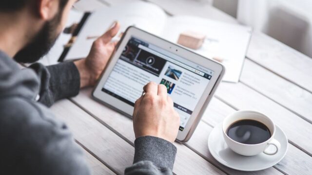 Man reading long-form content on a tablet at a white wooden table with a cup of coffee beside him