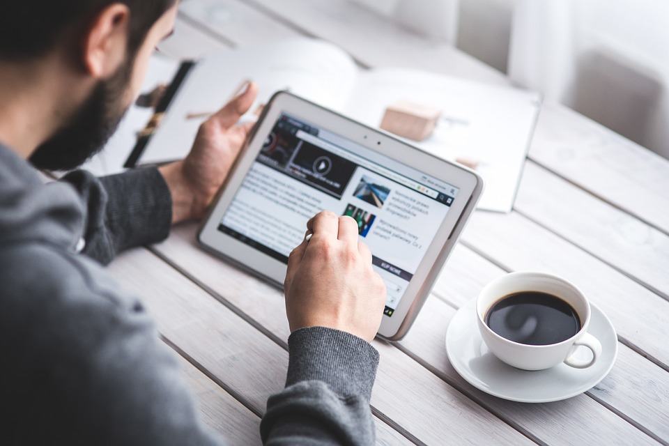 Man reading long-form content on a tablet at a white wooden table with a cup of coffee beside him